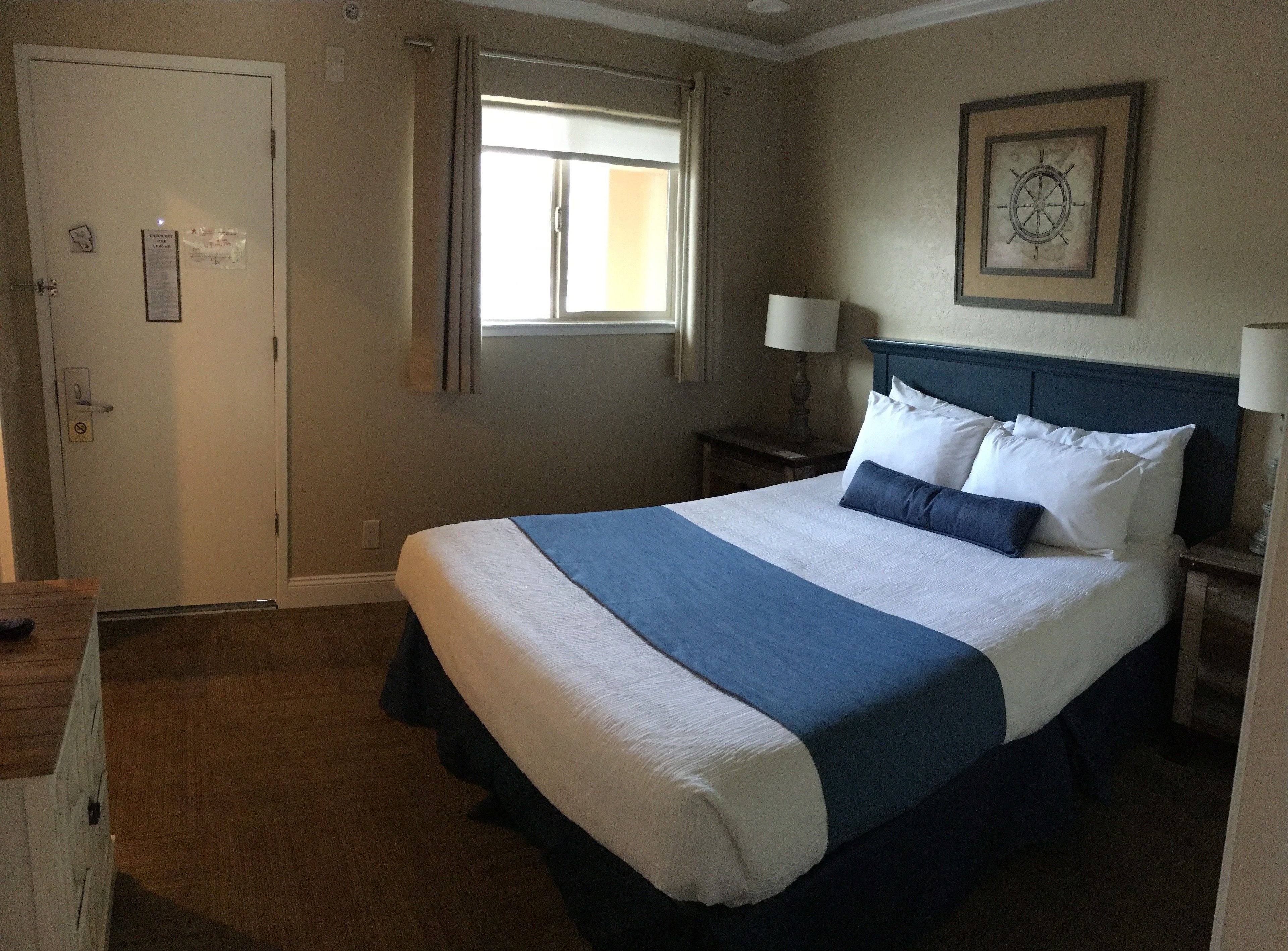 Habitación de hotel con una cama grande, una mesita de noche, una ventana con cortinas, una puerta con un cartel y un adorno de brújula en la pared.