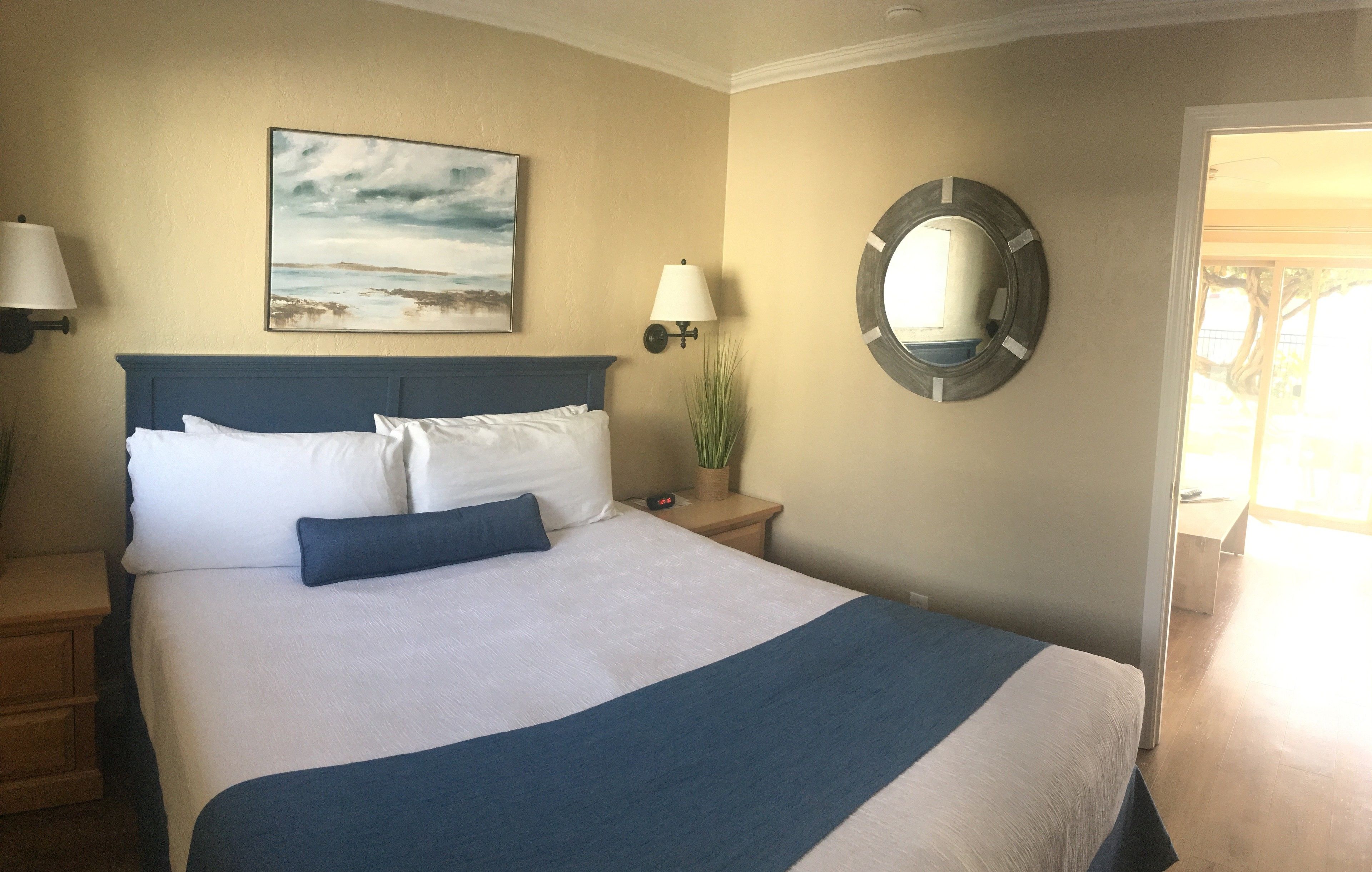 A neatly made bed with white and blue bedding, a painting on the wall, a round mirror, and a nightstand with a lamp.
