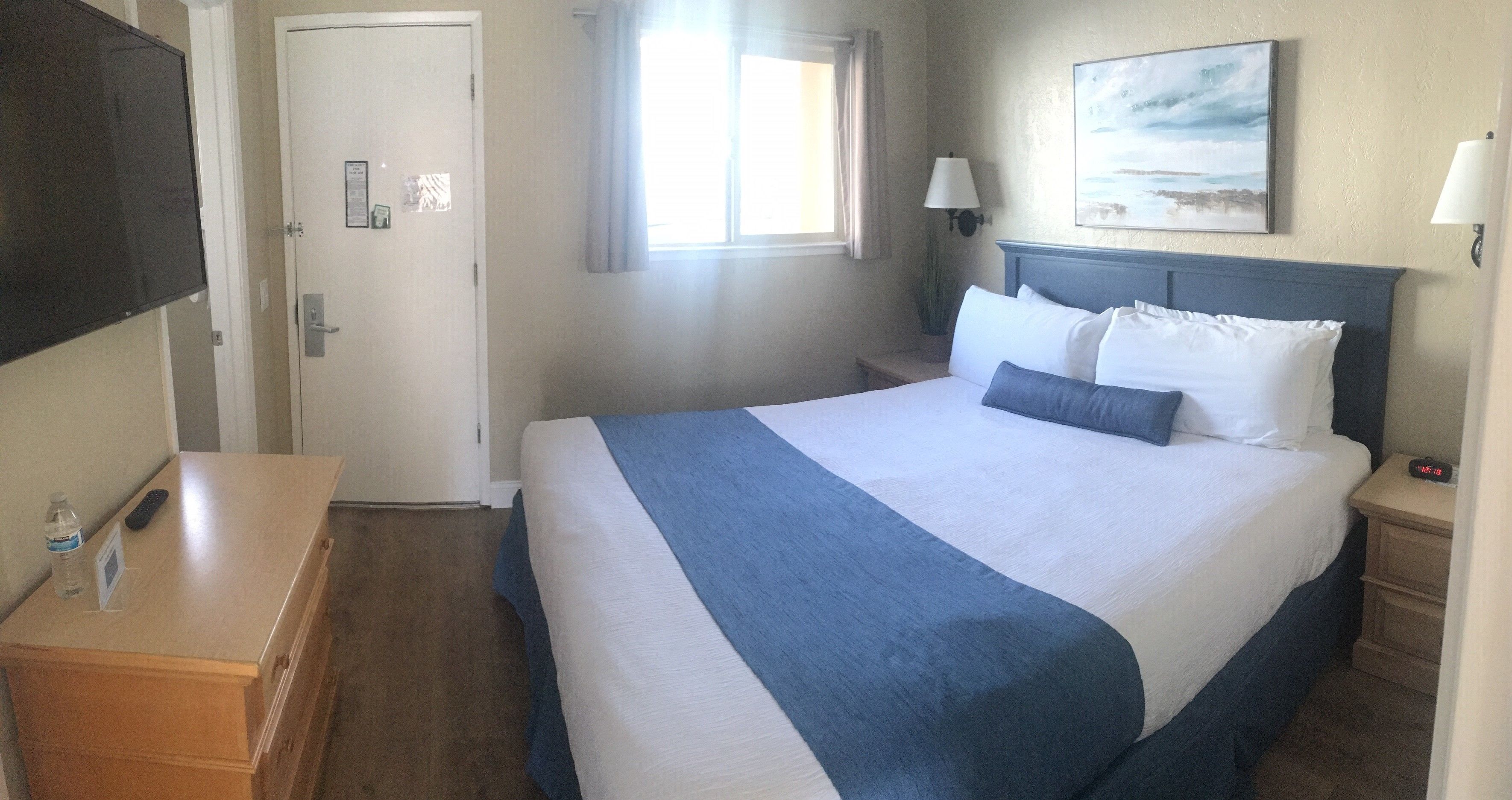 A hotel room with a large bed, a bedside table, a nightstand, a TV, a window with curtains, and a painting on the wall.