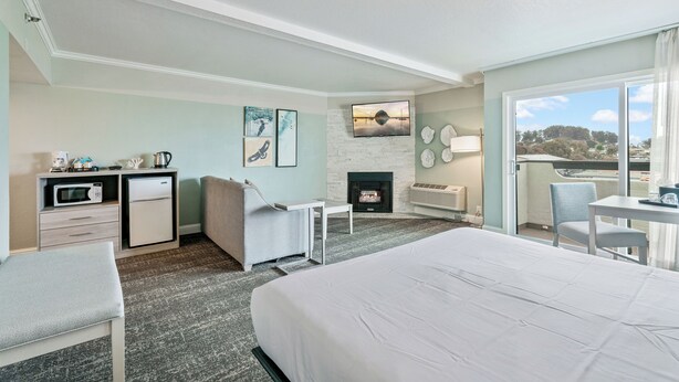 A hotel room with a bed, a desk with a microwave, a small fridge, a seating area with a chair, a fireplace, and a large window with a view. - La Serena Inn (Morro Bay)