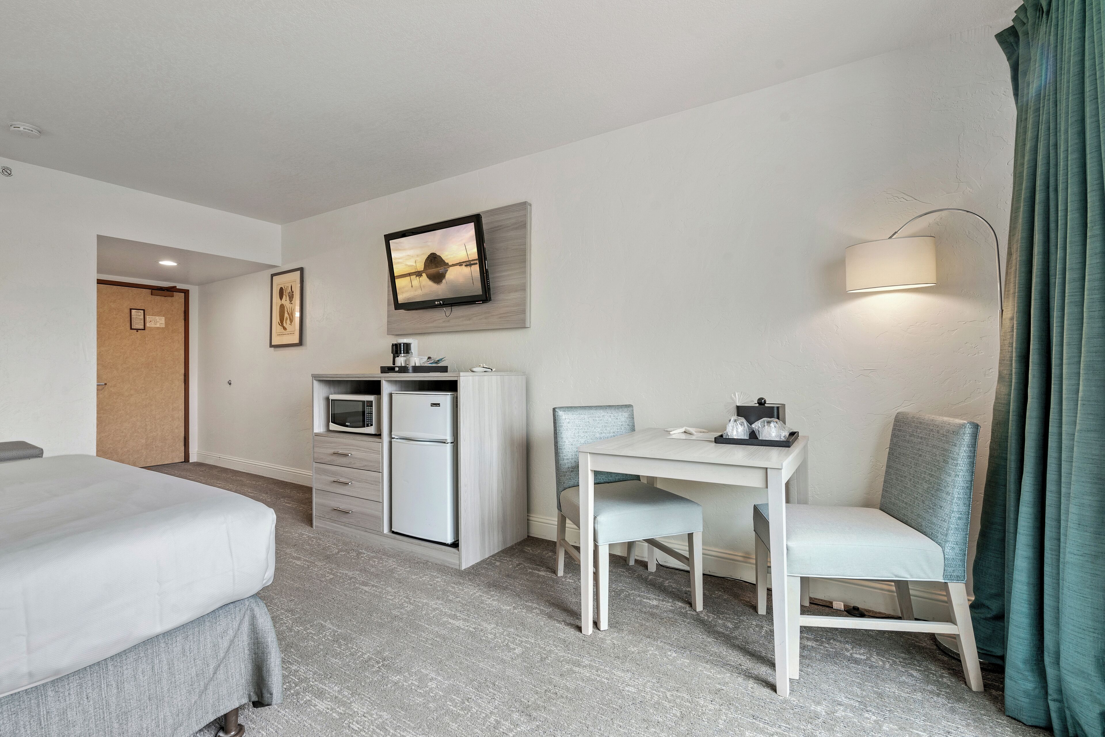 A hotel room with a bed, a desk with a chair, a television mounted on the wall, a microwave, and a small refrigerator.