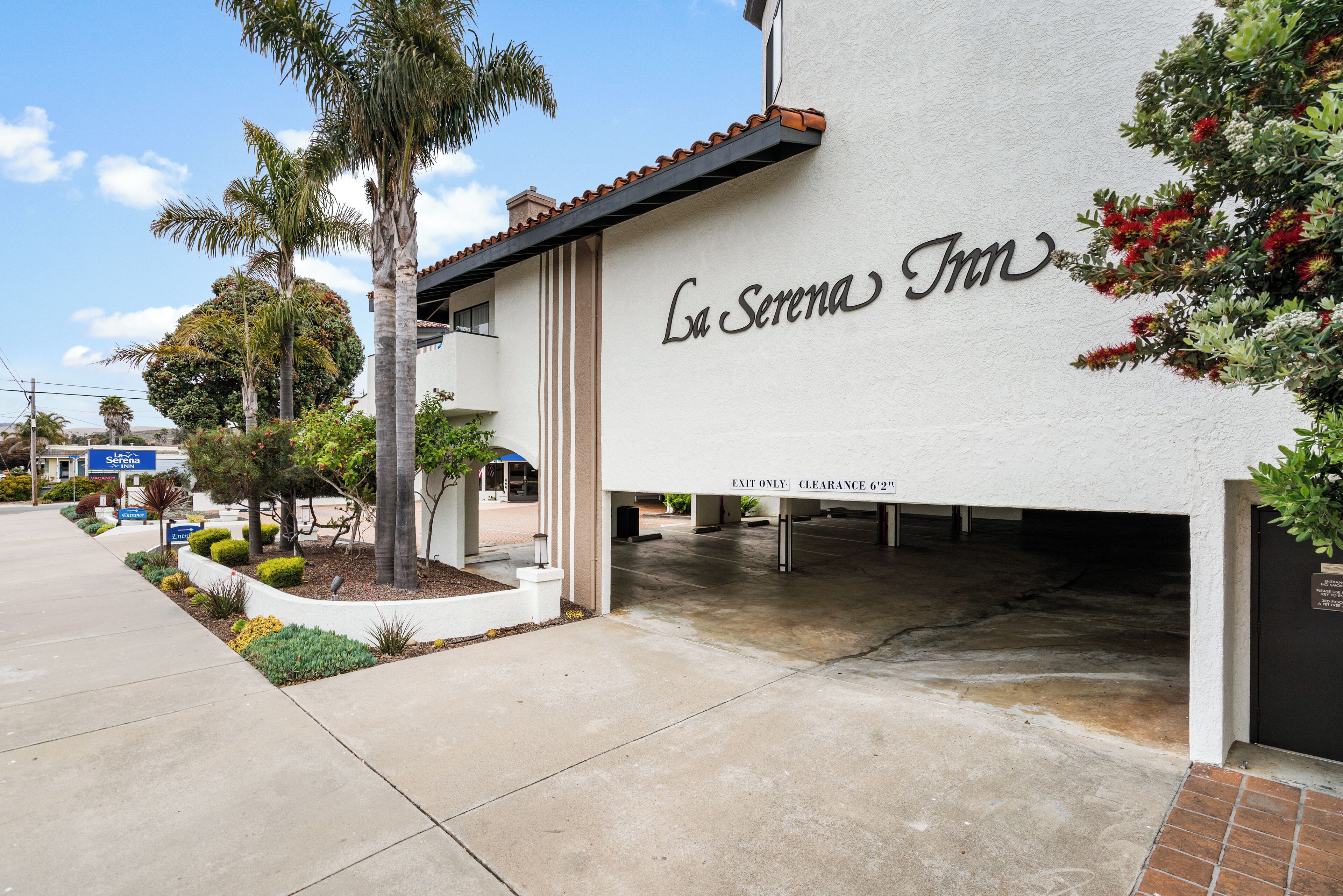 The exterior of La Serena Inn with a clear sign, a parking area, and a landscaped garden with palm trees and shrubs.