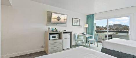 A hotel room with two beds, a desk with a microwave, a small refrigerator, a dining table with chairs, and a balcony with a view.