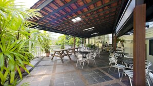 Un área de café al aire libre con mesas y sillas, plantas en macetas y una zona de estar cubierta.