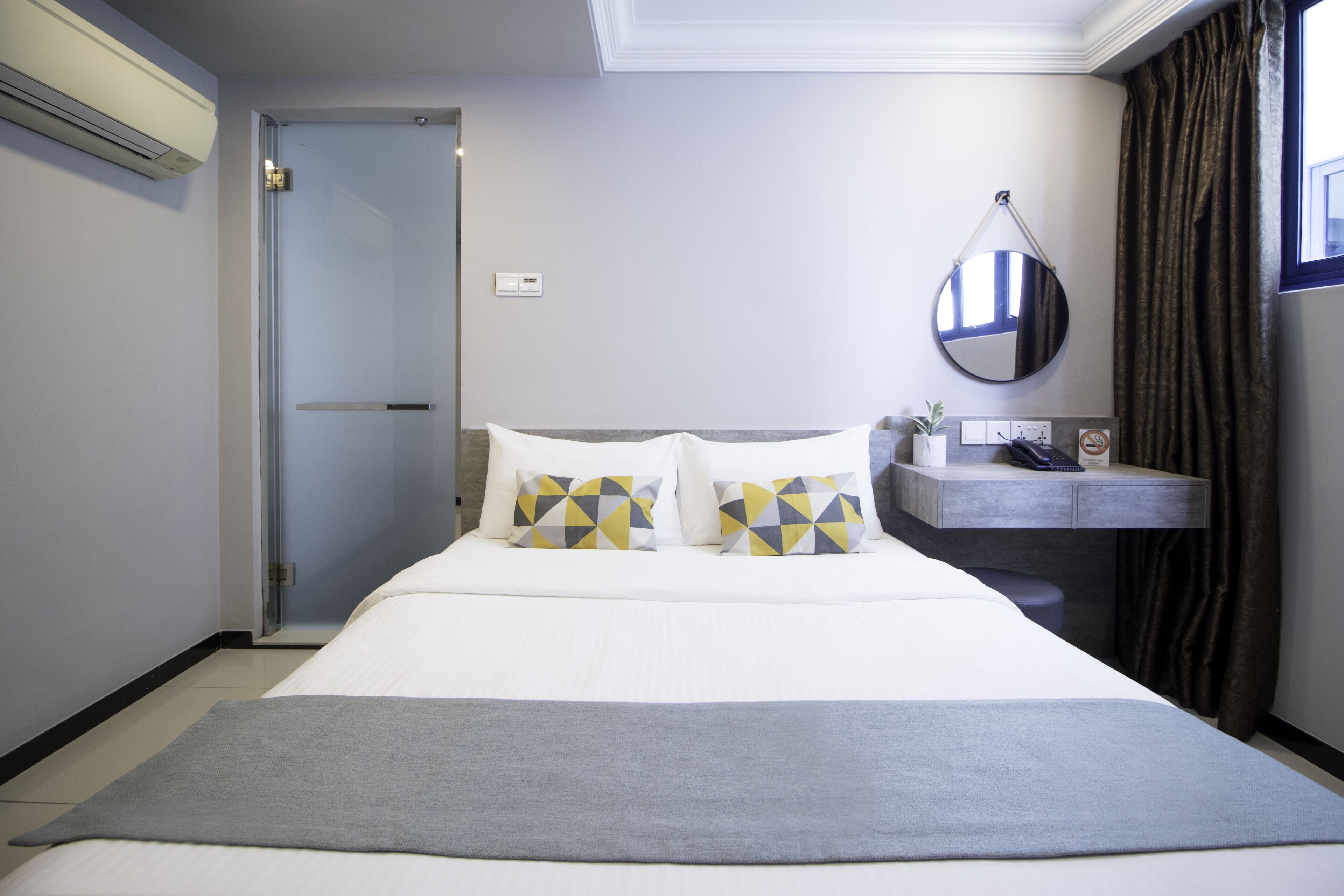 A hotel room with a bed, a bedside table, a mirror, and a window with curtains.