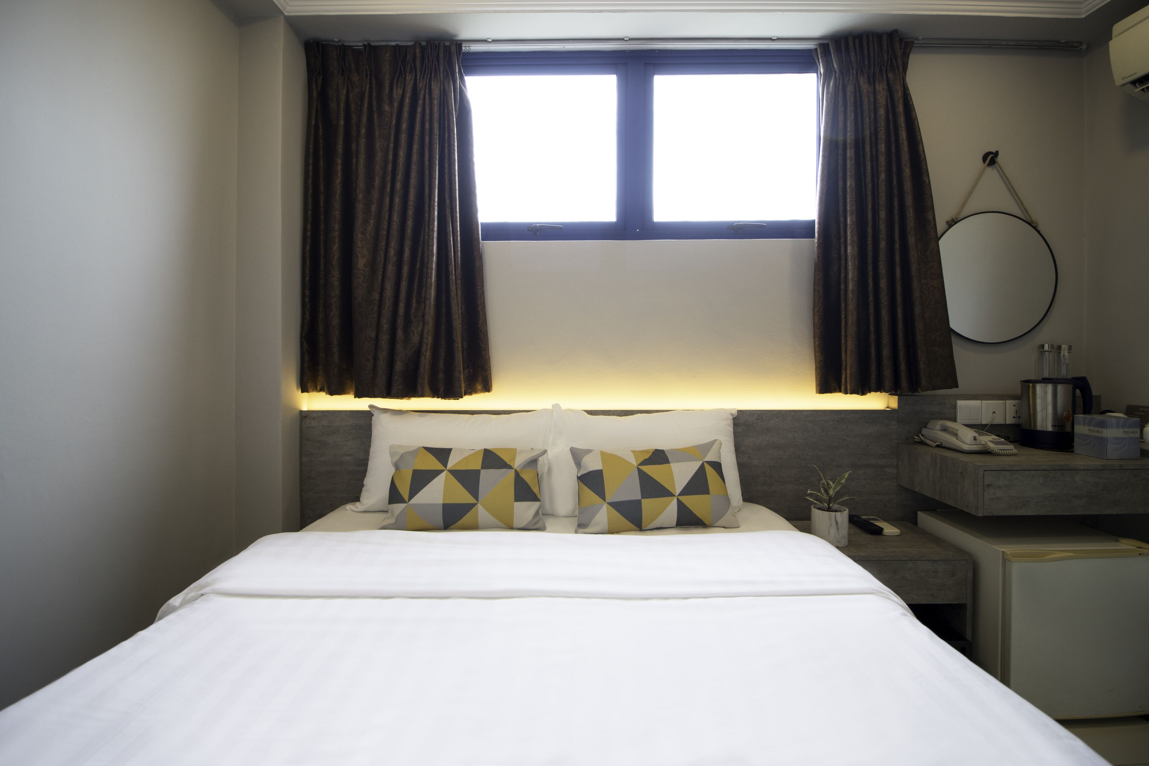 A hotel room with a bed, two pillows, a nightstand, a mirror, and a window with curtains.