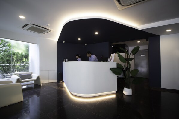 A modern reception area with a curved reception desk, two staff members, a white sofa, a large potted plant, and a view of greenery outside. - Harbour Ville Hotel (Singapore)