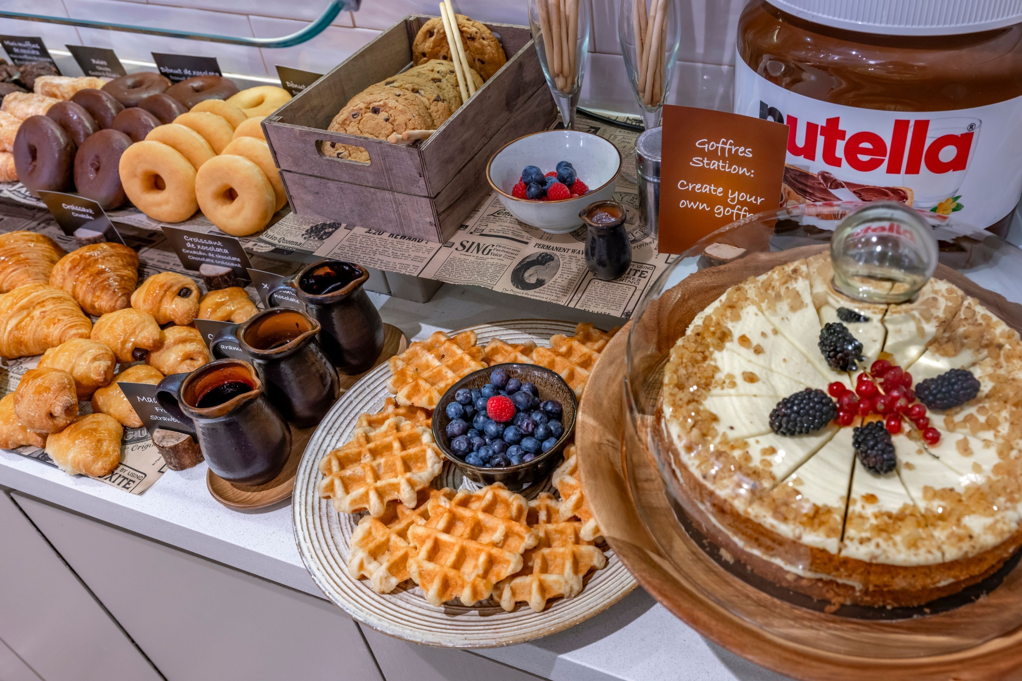 A variety of pastries, including croissants and waffles, with a Nutella jar and a bowl of berries.