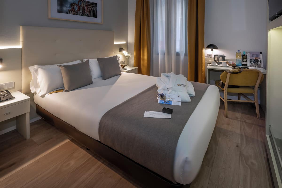 A hotel room with a large bed, a desk, a chair, and a nightstand with a phone and a lamp.