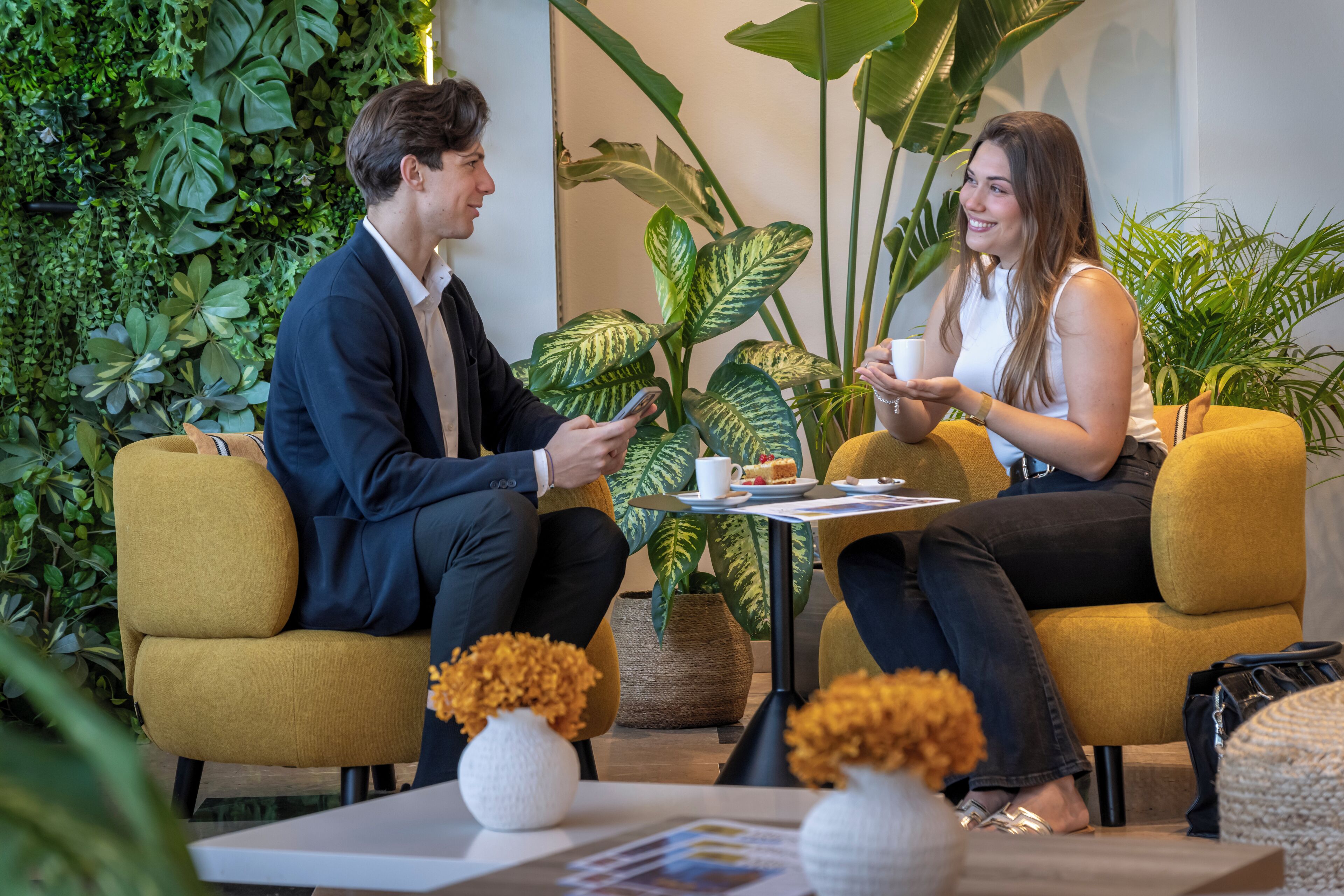 Two people seated on yellow chairs in a room with green plants and a white table.