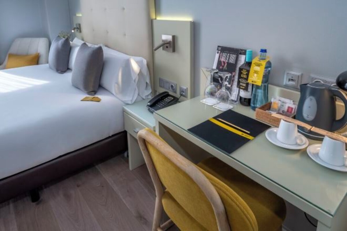 Quarto de hotel com uma cama, uma escrivaninha com cadeira, uma cafeteira e um telefone.