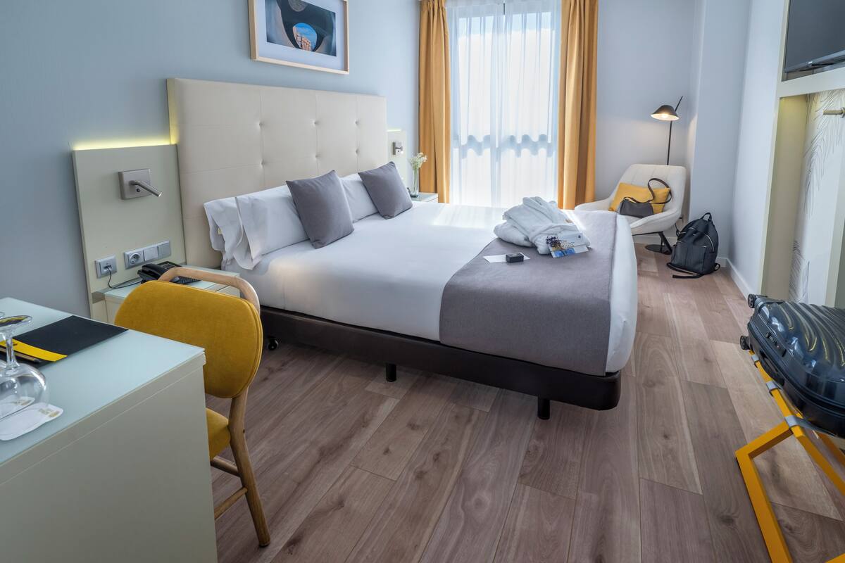 A modern hotel room with a large bed, a desk, a chair, a suitcase, and a window with curtains.