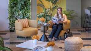 A woman sitting on a sofa reading a magazine in a lobby with a coffee table, white vases, and a potted plant.