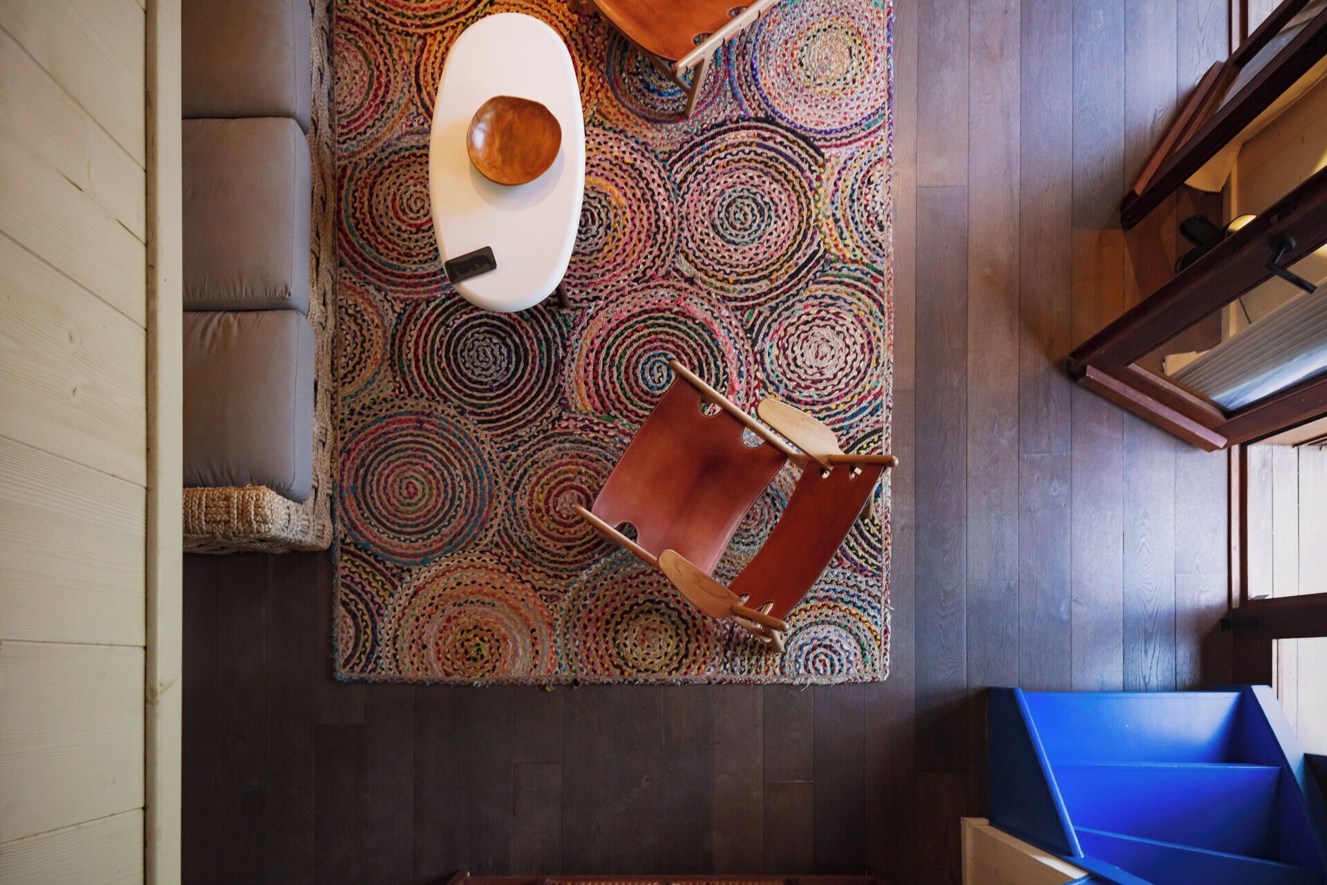 Een kamer met een donker houten vloer, een kleurrijk rond tapijt met een patroon, een bruine leren fauteuil, een houten tafel met een schaal, en een blauwe bank.