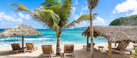 A beach with thatched umbrellas, wooden lounge chairs, and palm trees.