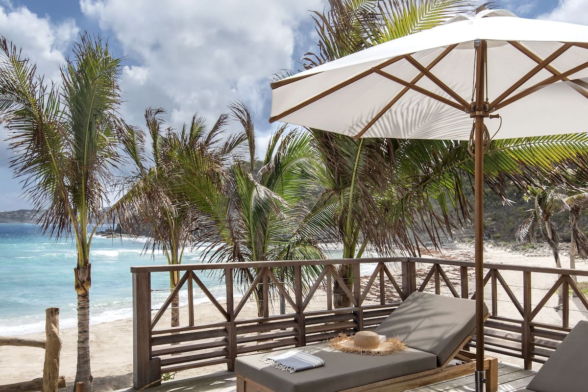 Une terrasse en bois avec des chaises longues et une table, donnant sur une plage bordée de palmiers et sur l’océan.