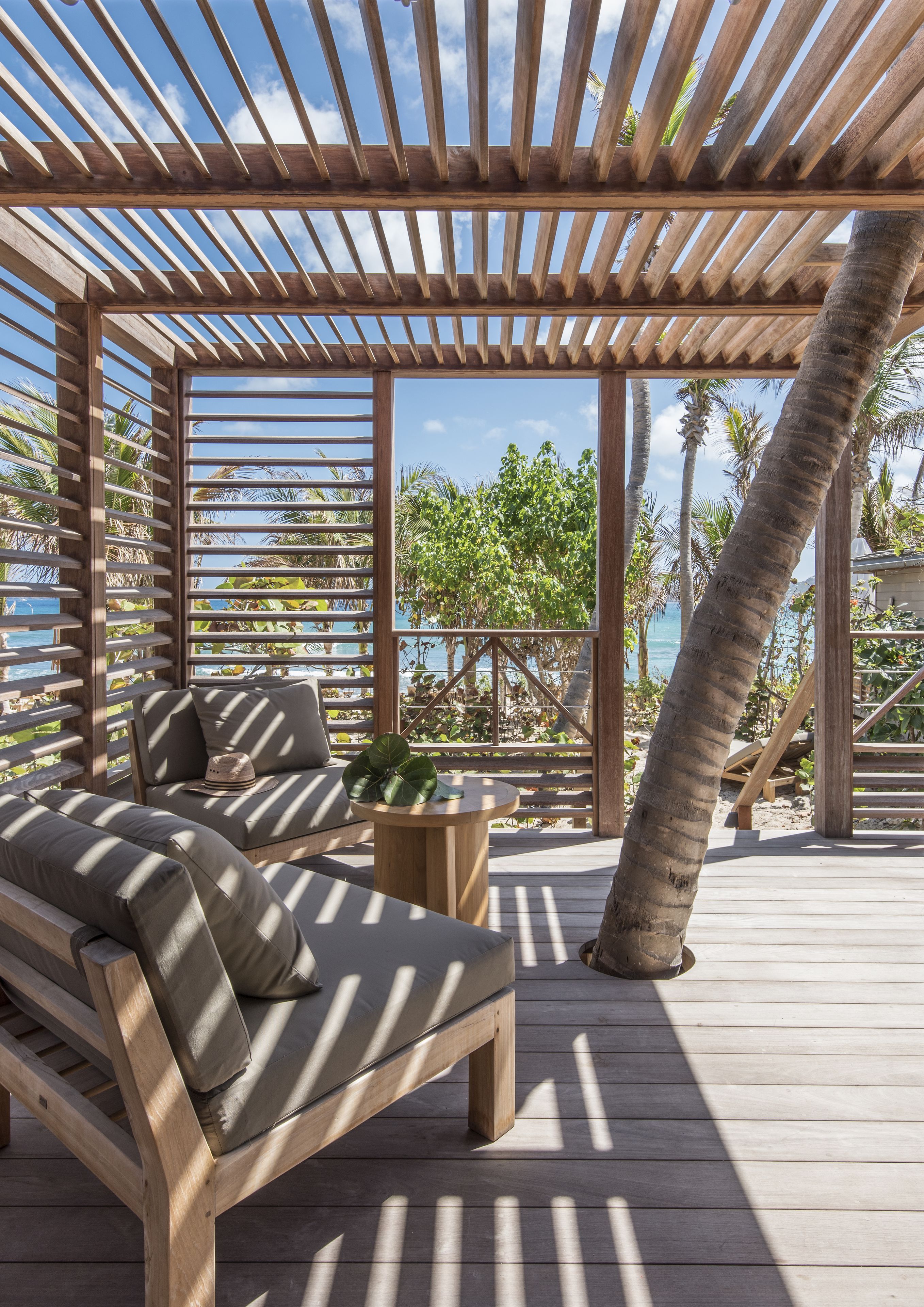 Eine Holzterrasse mit einer Couch und einem Tisch, mit Blick auf einen Strand mit Palmen und den Ozean.