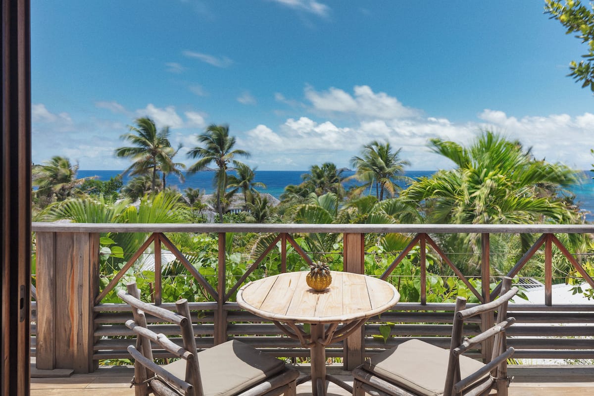 Une terrasse en bois avec une table ronde et des chaises, donnant sur une plage bordée de palmiers et sur l’océan.