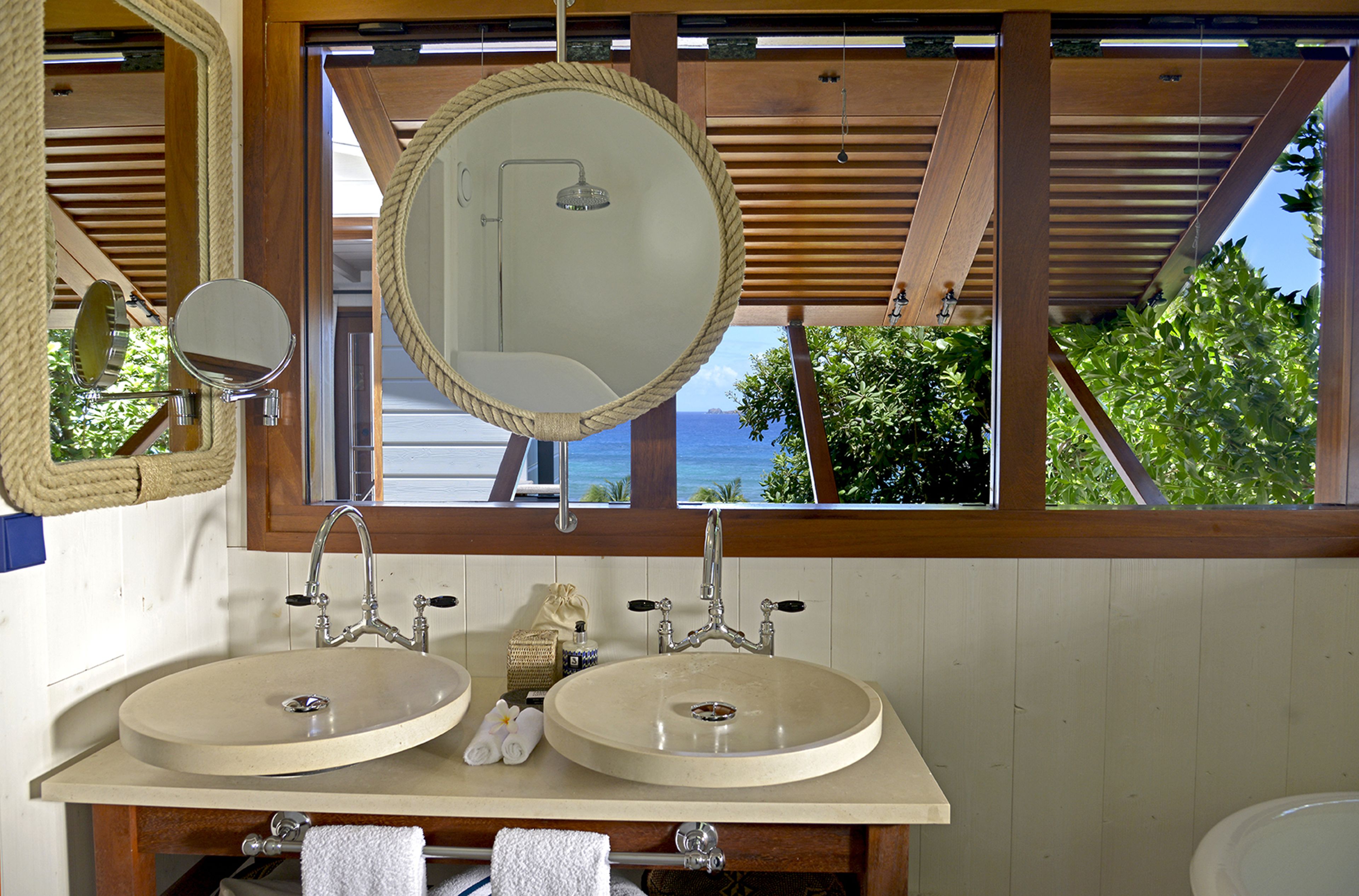 A bathroom with two sinks, a round mirror, and a view of the ocean.