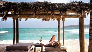 A beach scene with a wooden structure, a table with a drink, and a person lying on a lounge chair. - Hotel Manapany (St. Barthelemy)