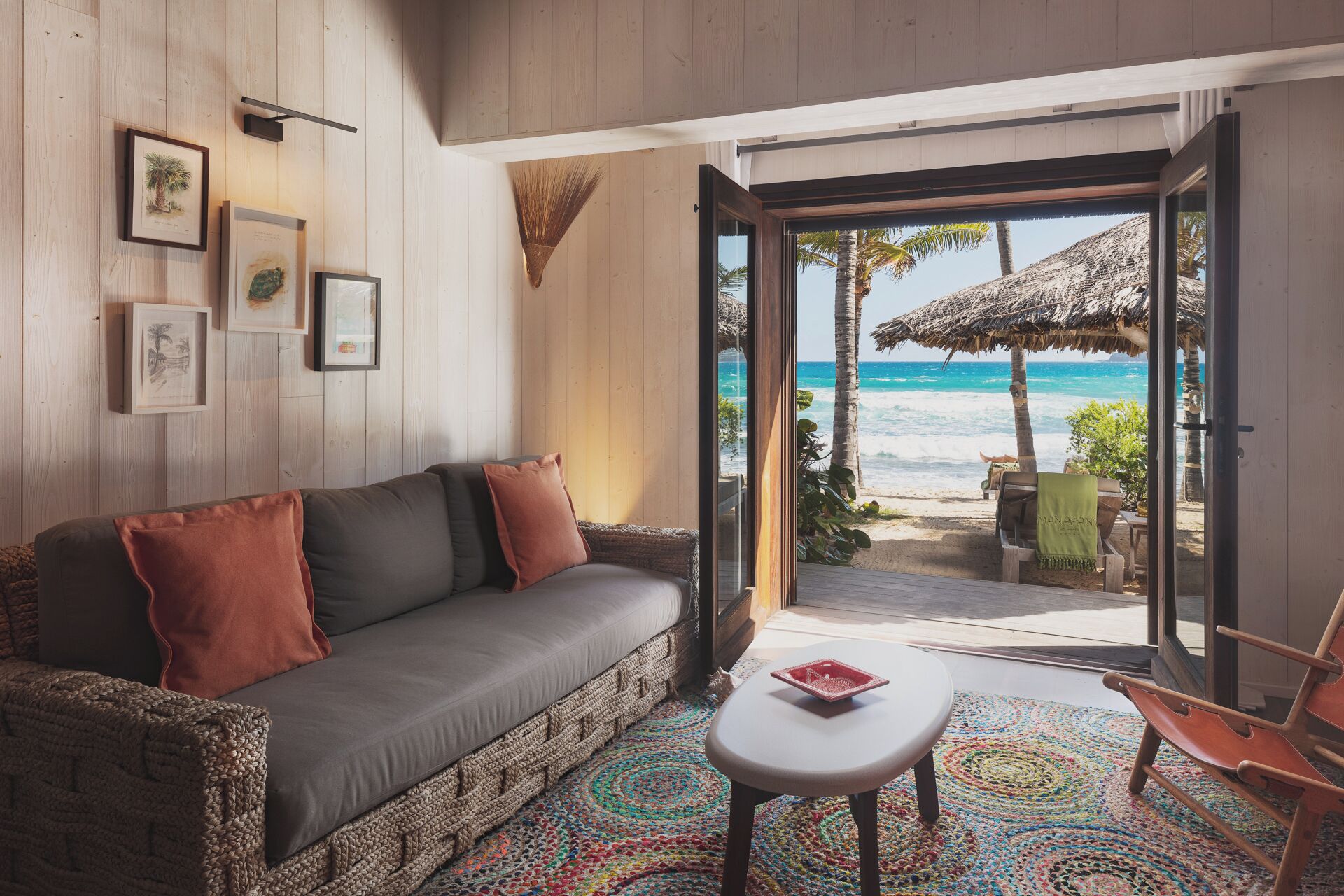A cozy room with a sofa, a small table, and a colorful rug. There are framed pictures on the wall and a view of the beach through an open door.