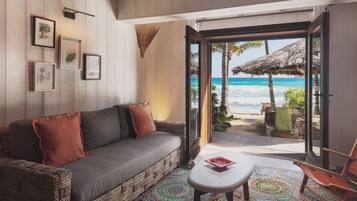 A cozy room with a sofa, a small table, and a colorful rug. There are framed pictures on the wall and a view of the beach through an open door.