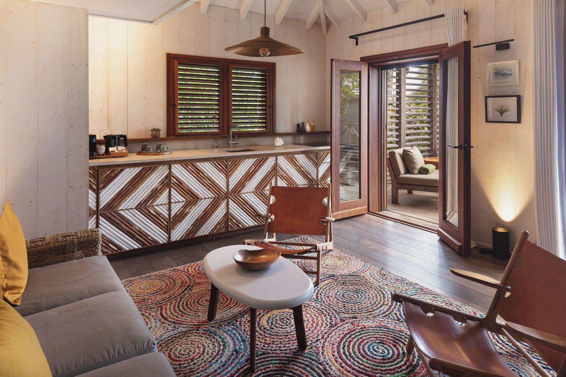 Un cuarto acogedor con un sofá, una silla, una mesita y una alfombra colorida. Cuenta con una zona de cocina con mostrador y una ventana con persianas.