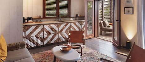 Un cuarto acogedor con un sofá, una silla, una mesita y una alfombra colorida. Cuenta con una zona de cocina con mostrador y una ventana con persianas.