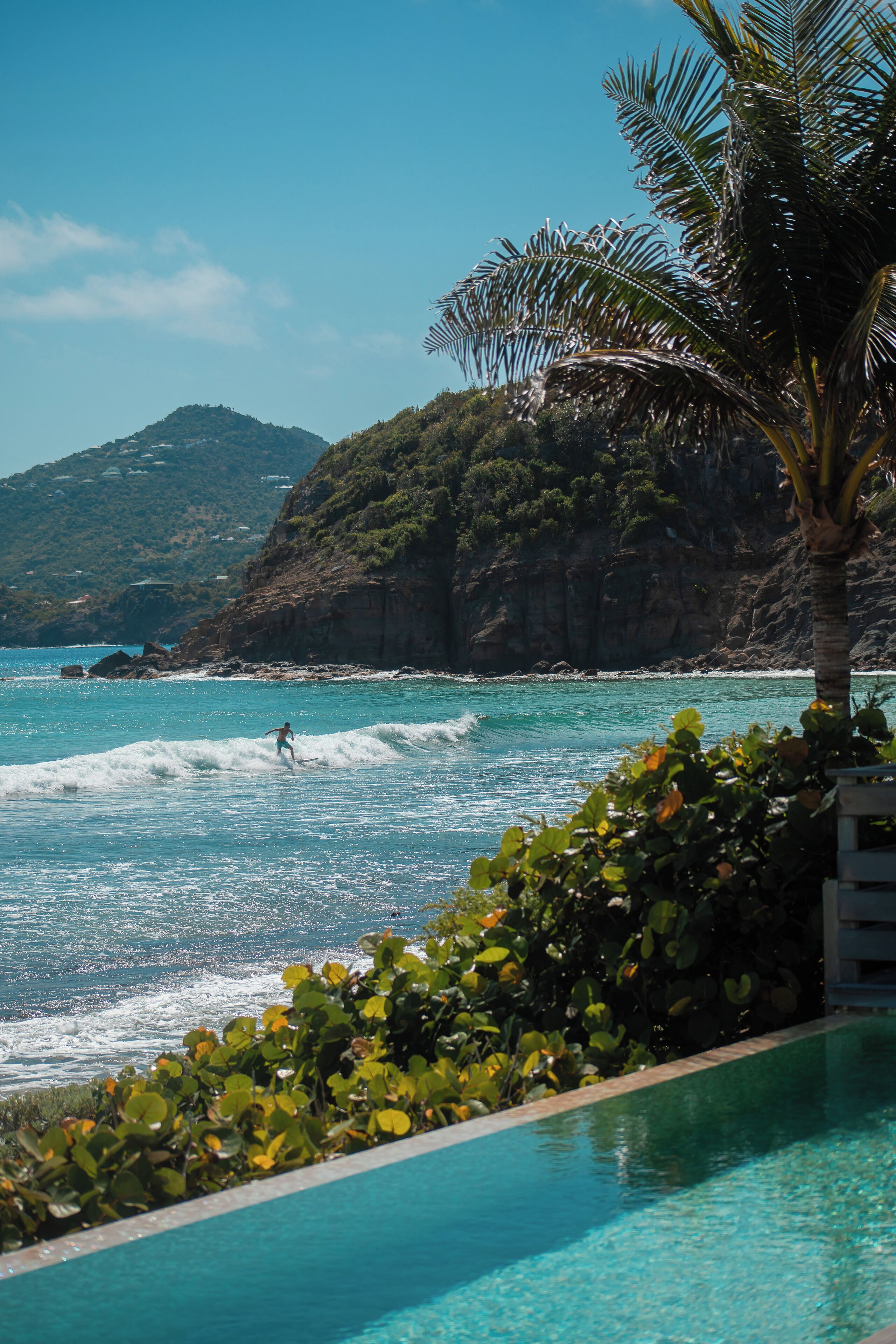 A poolside view with clear blue water, a person surfing, and a mountainous backdrop.