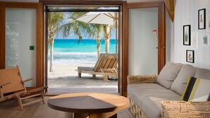 A room with a view of a beach, a wooden chair, a sofa, and a round table. - Hotel Manapany (St. Barthelemy)