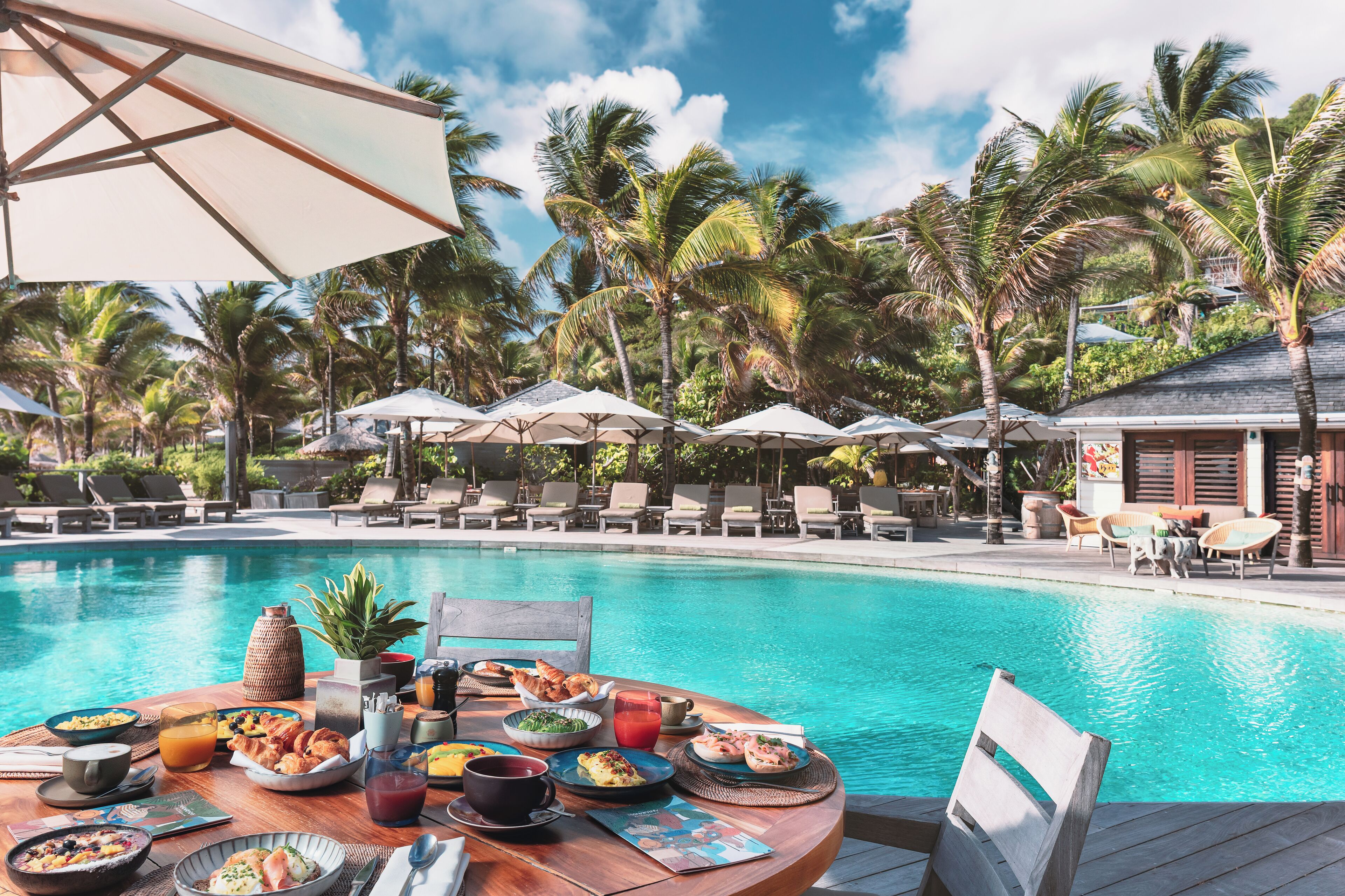 Un área de comedor junto a la piscina con una mesa dispuesta para una comida, rodeada de sillones de descanso y sombrillas, con palmeras al fondo.
