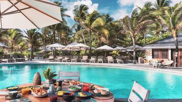 Un área de comedor junto a la piscina con una mesa dispuesta para una comida, rodeada de sillones de descanso y sombrillas, con palmeras al fondo.