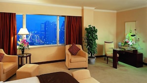 Une chambre d’hôtel avec une grande fenêtre, deux fauteuils, un canapé, un bureau, une lampe, une plante et un vase rempli de fleurs.