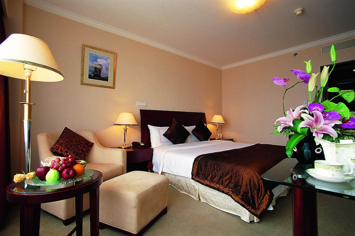 Une chambre d’hôtel avec un lit, des lampes de chevet, une petite table avec des fruits, une chaise et un vase rempli de fleurs.