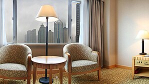 Une chambre d’hôtel avec deux fauteuils à motifs, une petite table ronde, une lampe et une vue sur les bâtiments de la ville par la fenêtre.