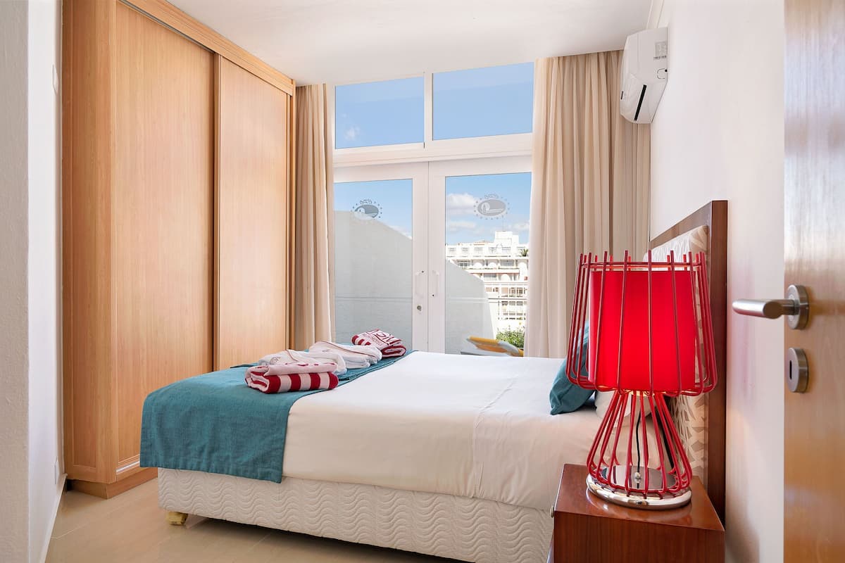 Una habitación de hotel con una cama, una lámpara roja, un armario y una ventana con vistas a edificios.