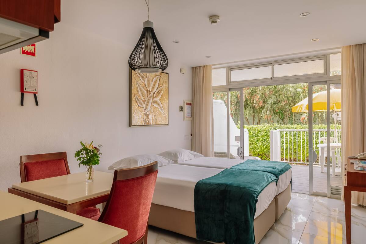 Habitación de hotel con cama, escritorio con silla, una mesita con un jarrón de flores y un balcón con sombrilla amarilla.