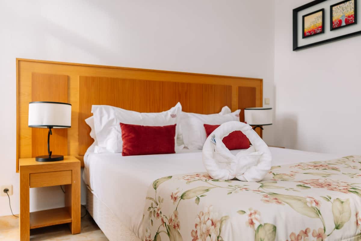 Una cama bien hecha con una colcha estampada con motivos florales, almohadas rojas y una toalla blanca sobre la cama.