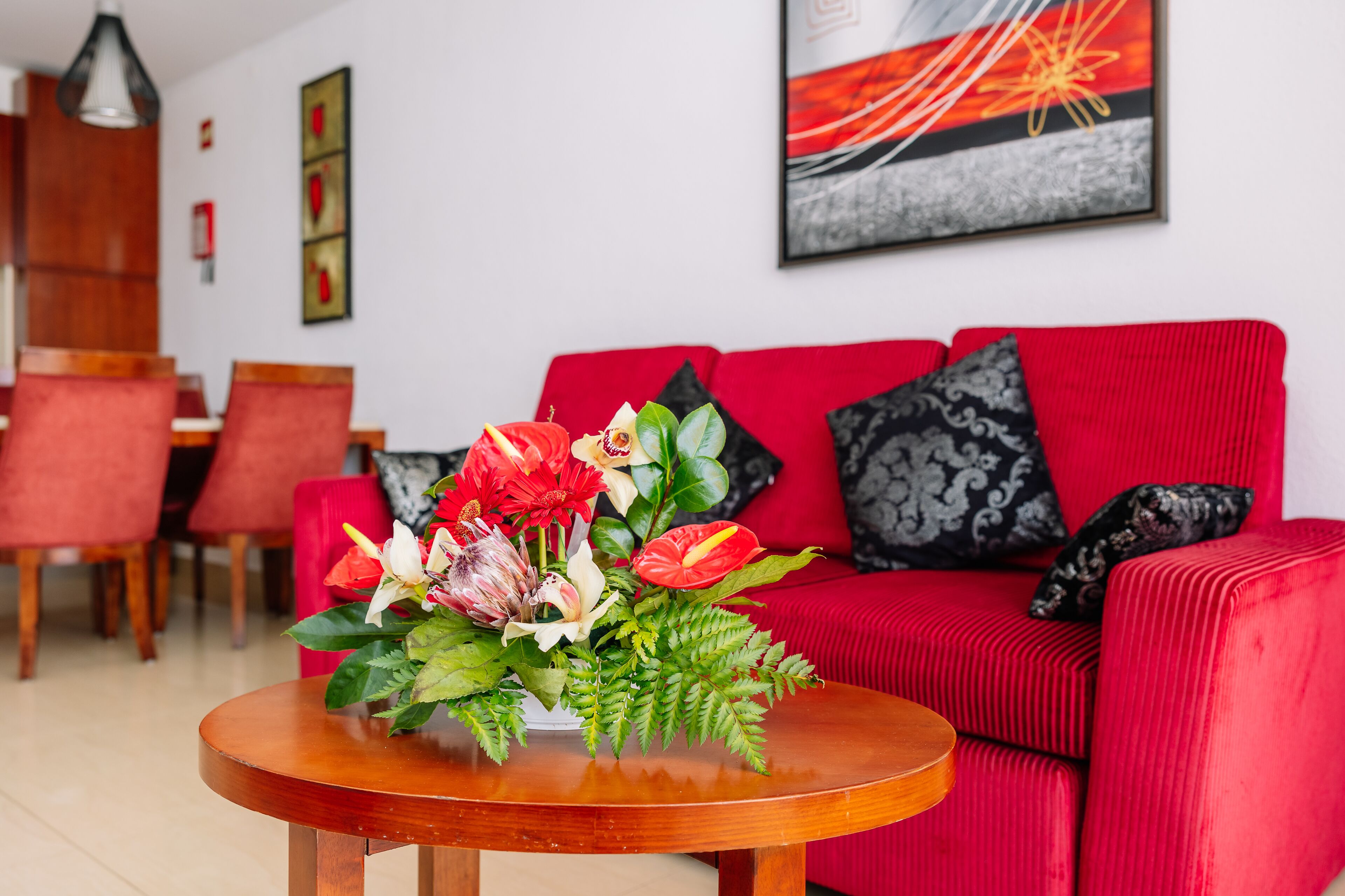 Una sala de estar moderna con un sofá rojo, una mesa de centro de madera con un arreglo floral, y un área de comedor al fondo.