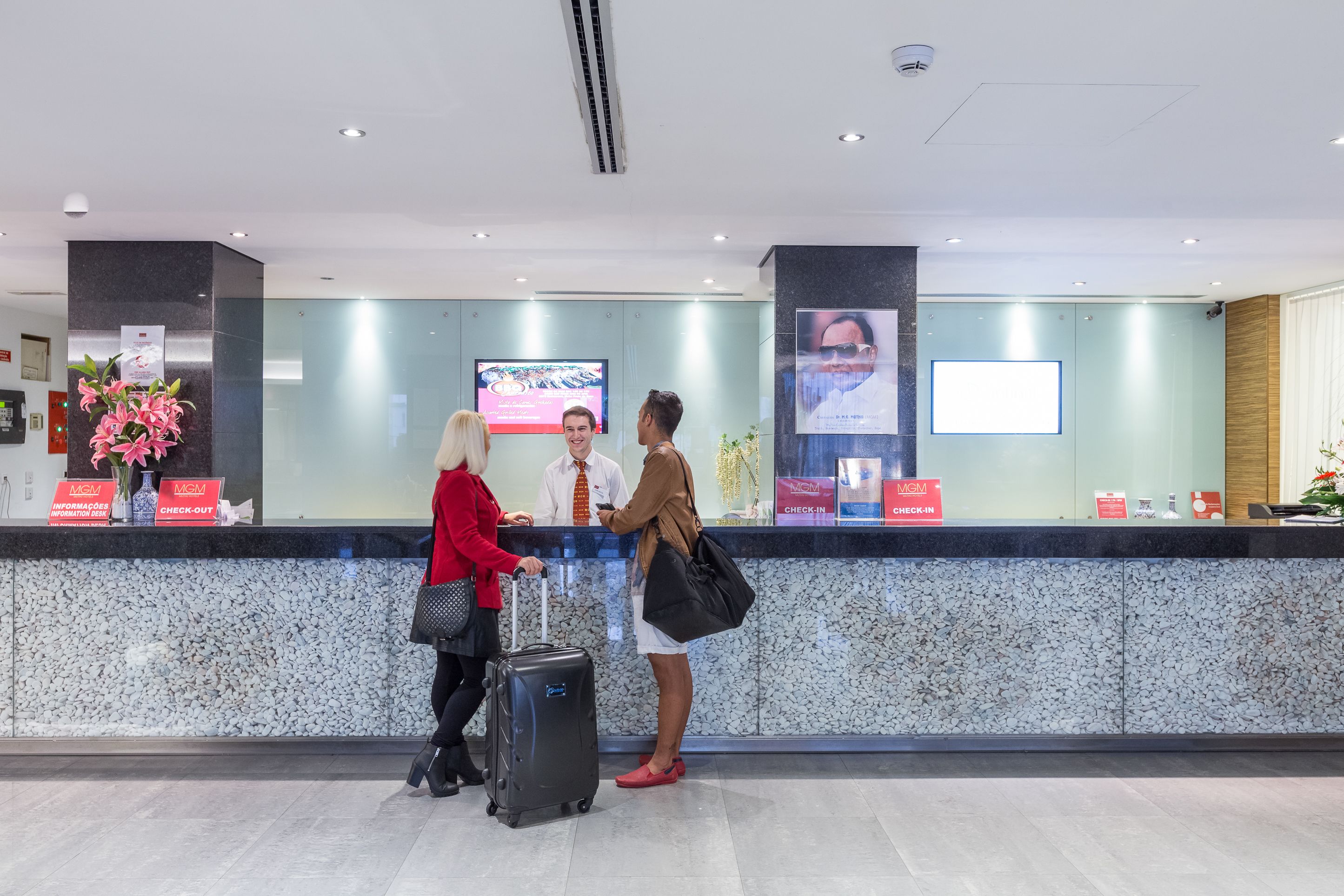 A hotel reception area with a check-in counter, a digital screen, and a large poster.