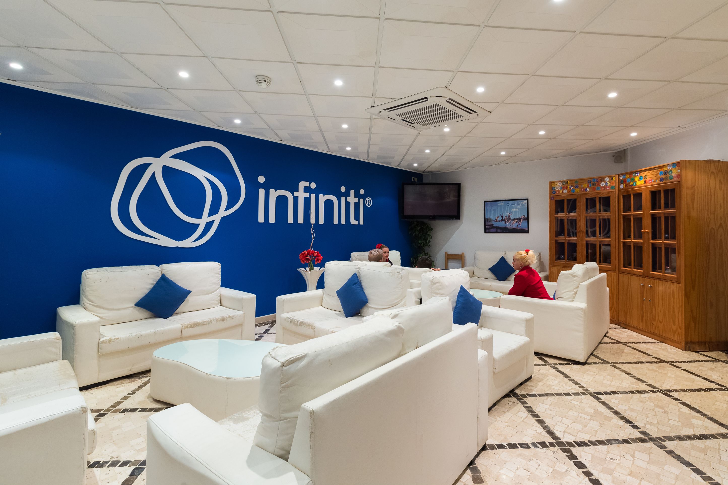 Un vestíbulo con sofás blancos, una pared azul con el logo 'infiniti', un mueble de madera y un suelo de baldosas.