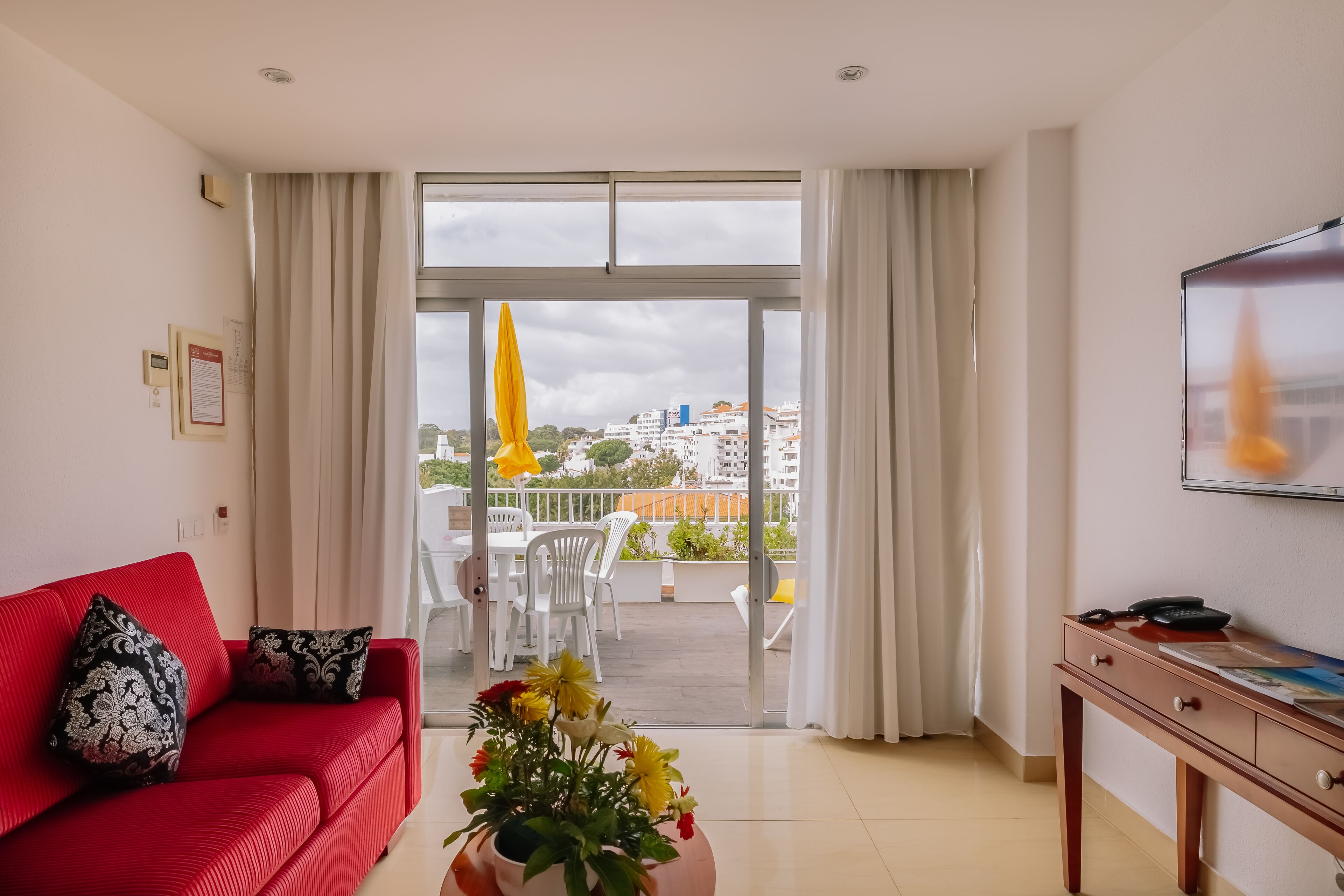 Una habitación de hotel moderna con un sofá rojo, una mesita de madera y un balcón con una sombrilla amarilla.