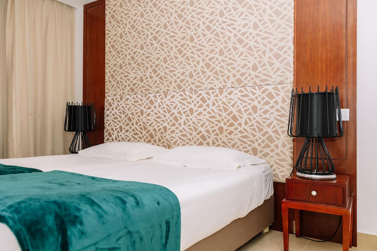 Una cama bien hecha con sábanas blancas, un plaid color turquesa y una mesita de noche de madera con una lámpara.