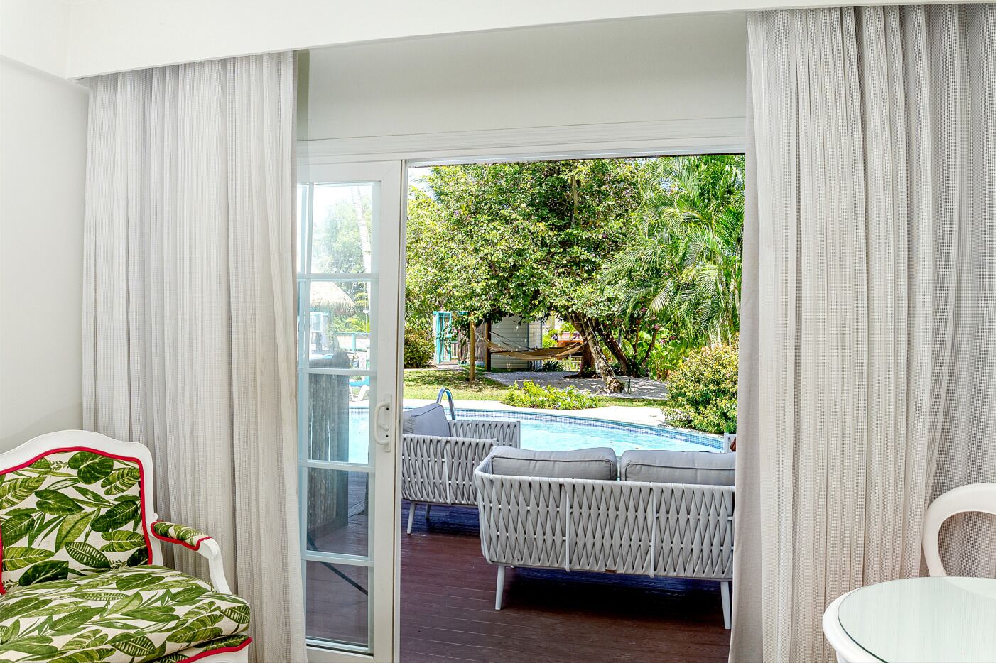 Una habitación con puerta de vidrio corredera que da a una zona de piscina, muebles de mimbre y vistas a un jardín exuberante.