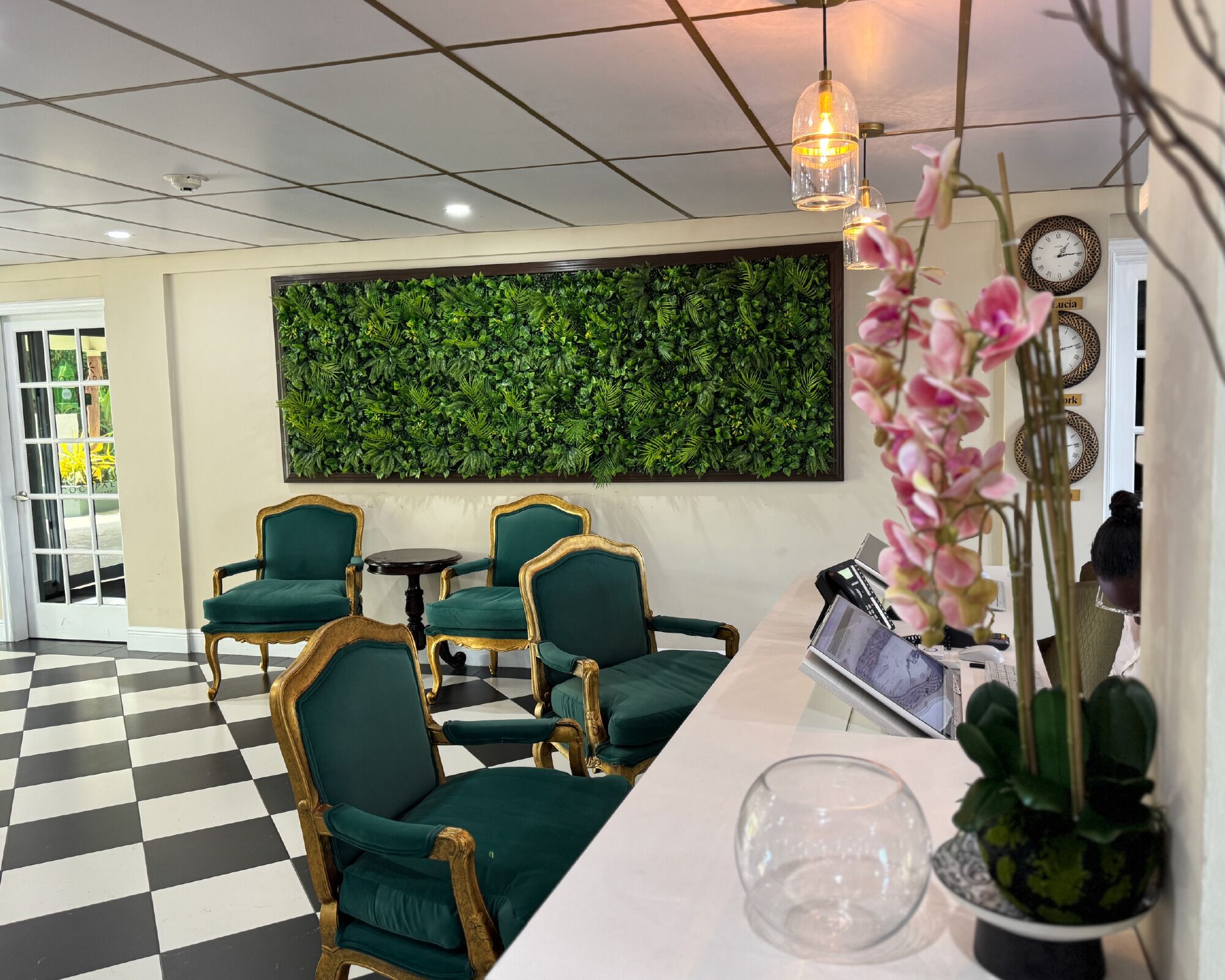 Una zona de recepción con piso a cuadros, sillas tapizadas de verde, un conjunto de follaje verde montado en la pared, un reloj y una planta en maceta.