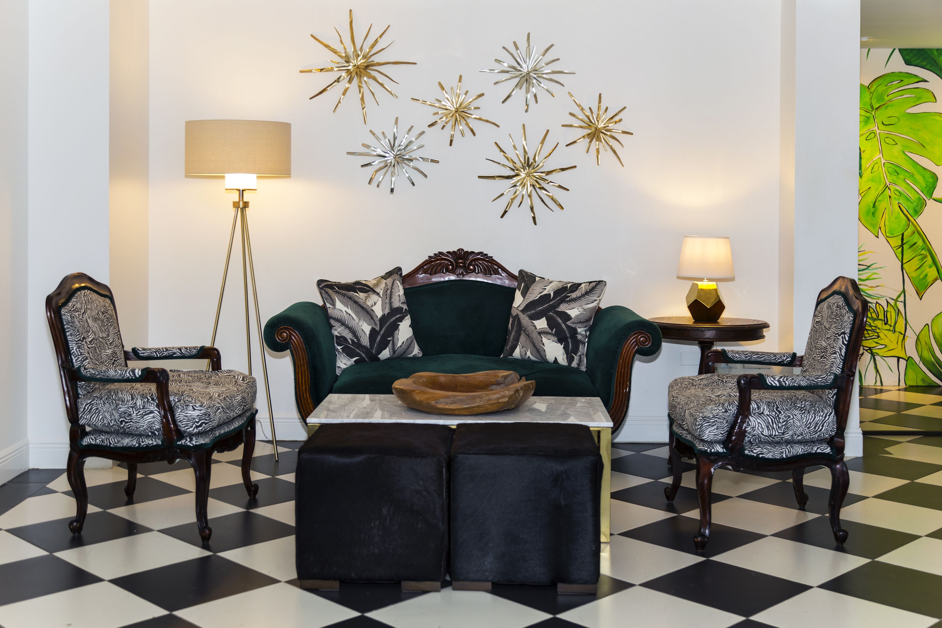 Una habitación con piso a cuadros, un sofá verde con cojines estampados, dos sillones estampados, una mesita redonda con un cuenco y decoraciones en la pared con diseños de estrellas y hojas.