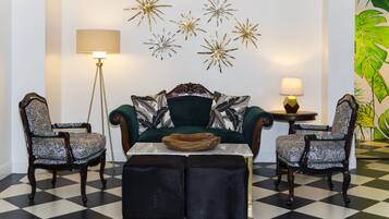 Una habitación con piso a cuadros, un sofá verde con cojines estampados, dos sillones estampados, una mesita redonda con un cuenco y decoraciones en la pared con diseños de estrellas y hojas.
