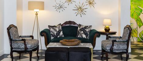 Una habitación con piso a cuadros, un sofá verde con cojines estampados, dos sillones estampados, una mesita redonda con un cuenco y decoraciones en la pared con diseños de estrellas y hojas.