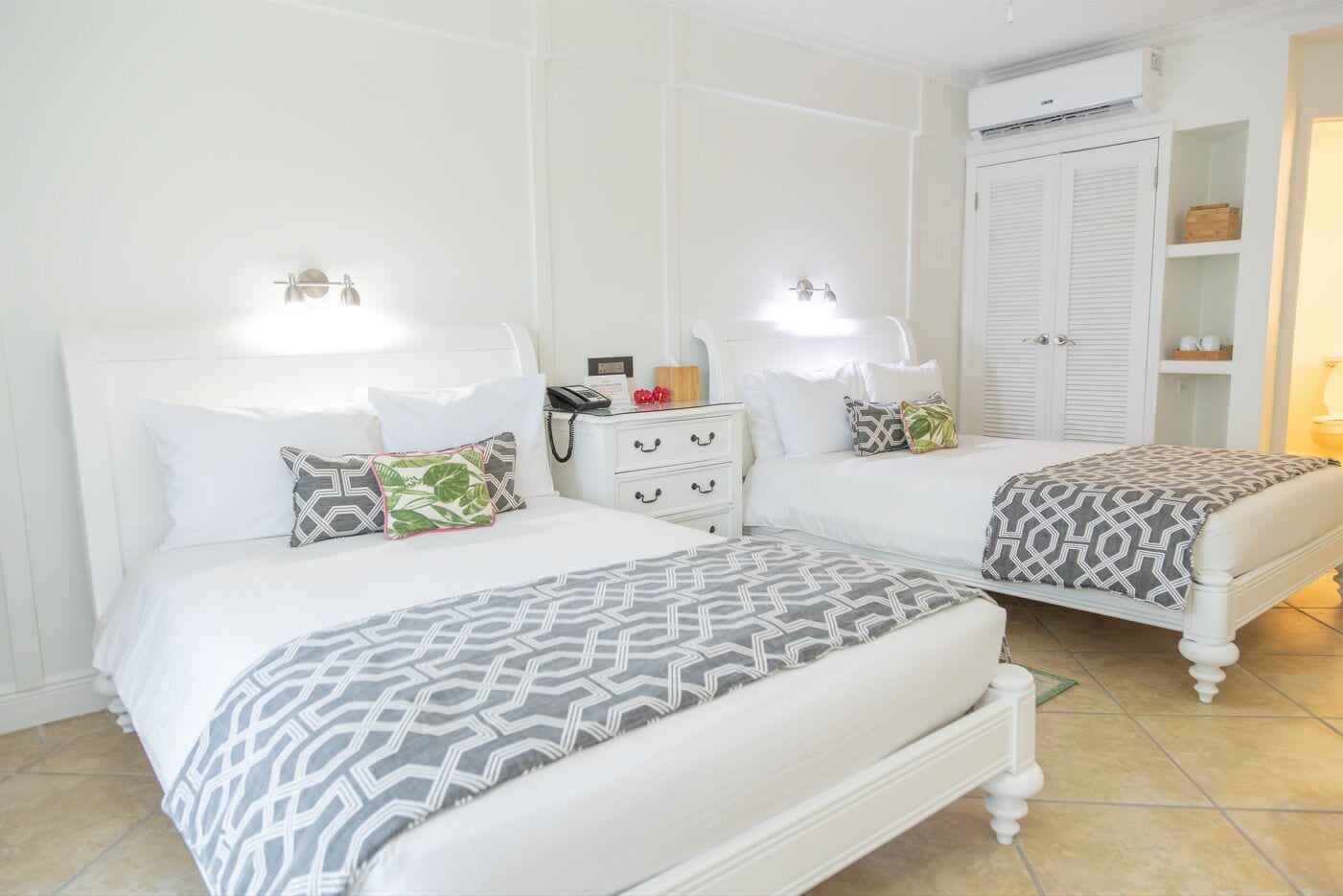 Une chambre d’hôtel avec deux lits, des murs blancs, une armoire blanche et un appareil de climatisation.