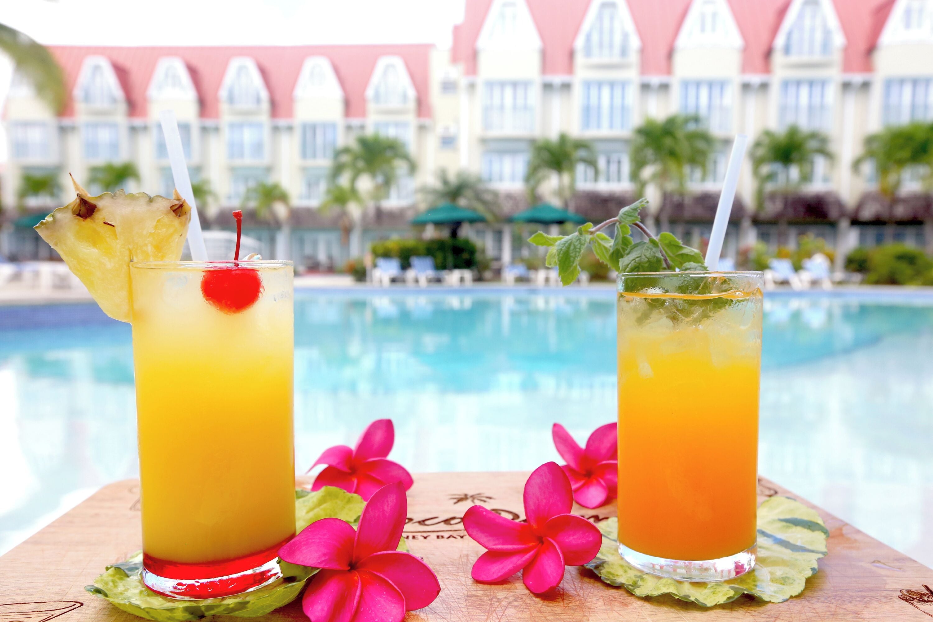 Two cocktails with a tropical theme, a pool, and a resort building in the background.